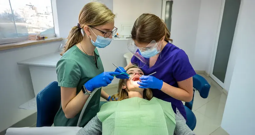 Two dental professionals in protective gear and masks performing a dental procedure on a patient in a modern dental office, showcasing teamwork and hygiene practices.